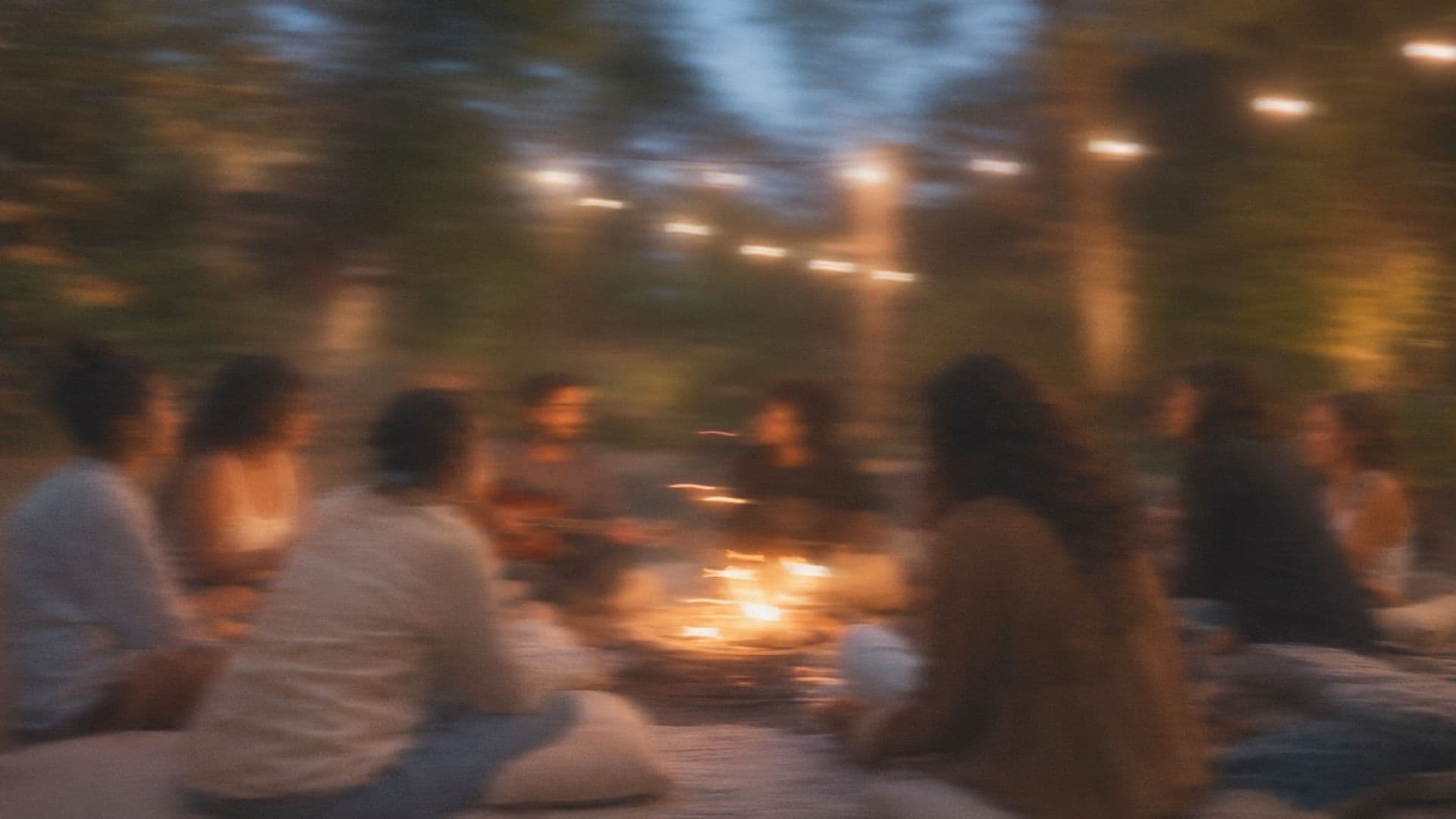 A community gathered in a circle around a fire, under string lights